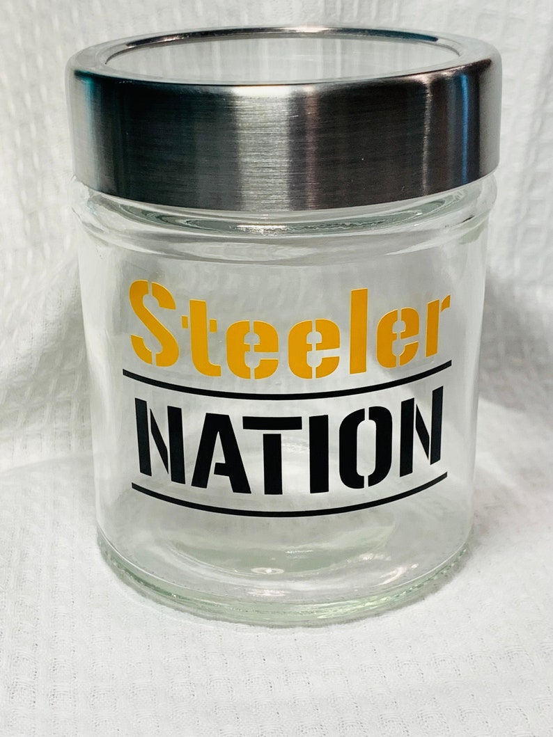 May include: A clear glass jar with a silver lid. The jar has a yellow and black decal that says "Steeler Nation".