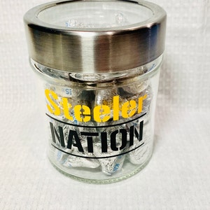 May include: A glass jar with a silver lid filled with foil-wrapped chocolate kisses. The jar has a black and yellow sticker that says "Steelers Nation".