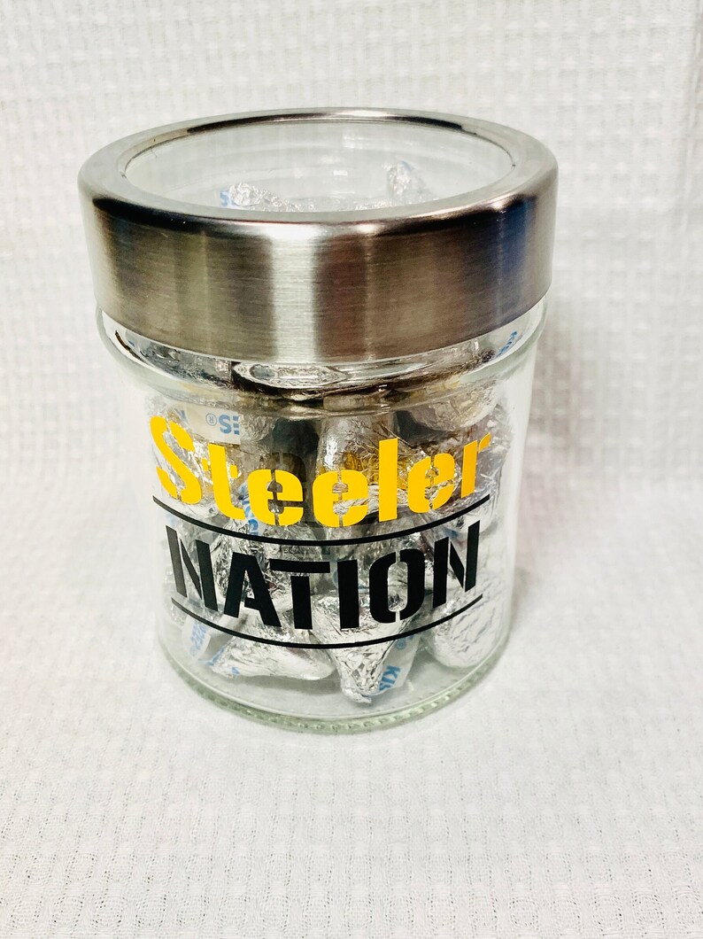 May include: A glass jar with a silver lid filled with foil-wrapped chocolate kisses. The jar has a black and yellow sticker that says "Steelers Nation".