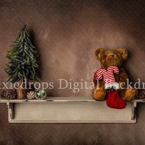 May include: A brown teddy bear wearing a red and white striped stocking sits on a white shelf with a small green Christmas tree and pine cones. The shelf is against a brown textured background.
