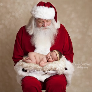 May include: Santa Claus in a red suit and white beard holds a sleeping baby wearing a gold headband. The baby is lying on a white fluffy blanket. The background is a light brown with a bokeh effect.