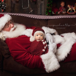 May include: Santa Claus in a red suit with white trim is sleeping on a brown couch. A baby wearing a red and white knitted hat and a red and white knitted blanket is being held by Santa.