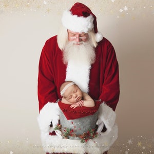 May include: Santa Claus in a red suit with white trim holds a baby in a metal bucket decorated with greenery. The baby is wearing a white headband and is asleep.