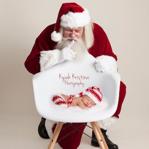 May include: Santa Claus in a red suit with a white beard and glasses is sitting in a white chair. A baby wearing a red and white striped outfit and a matching hat is sleeping in the chair. The text "Kyiah Kristina Photography" is printed on the chair.