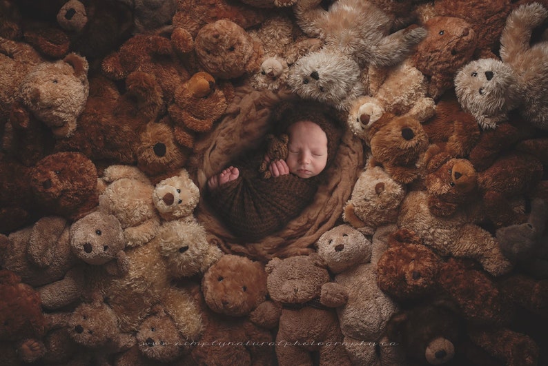 Puede incluir: Un beb&eacute; reci&eacute;n nacido envuelto en una manta marr&oacute;n, rodeado de numerosos osos de peluche. Los osos son de varios tonos marrones y beige, creando una escena c&aacute;lida y reconfortante. El beb&eacute; est&aacute; durmiendo pl&aacute;cidamente.