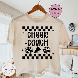 Può includere: T-shirt color crema con la scritta nera "Cheer Coach", pomponi e un megafono. Il design include un motivo a scacchi e stelle, perfetto per gli allenatori di cheerleading.