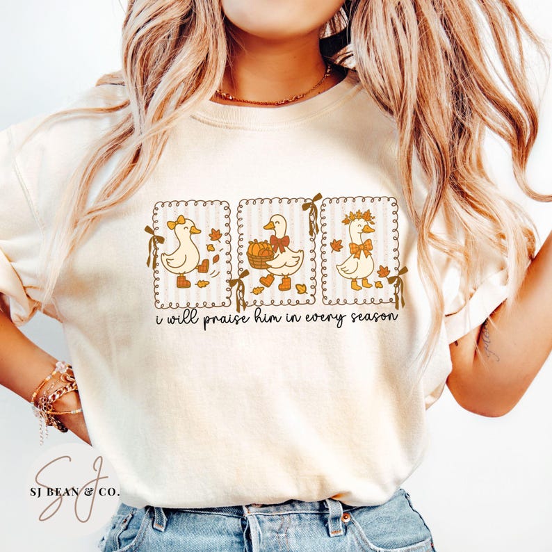 May include: Cream-colored sweatshirt featuring a whimsical design of three geese in decorative frames. Each goose is adorned with seasonal accessories, such as bows and a basket of pumpkins. The text "i will praise him in every season" is printed below the geese.