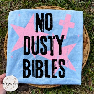 Peut inclure: Un t-shirt bleu clair avec le texte noir vieilli NO DUSTY BIBLES. Le motif comprend de grandes &eacute;toiles roses et un symbole de croix rose superpos&eacute;s derri&egrave;re le texte. Le t-shirt pr&eacute;sente un filigrane avec le texte SJ Bean & Co.