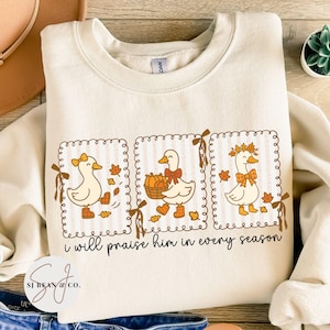 May include: Cream-colored sweatshirt featuring three cartoon ducks in decorative frames. Each duck is adorned with fall-themed accessories like bows, leaves, and pumpkins. The text "i will praise him in every season" is printed below the ducks.