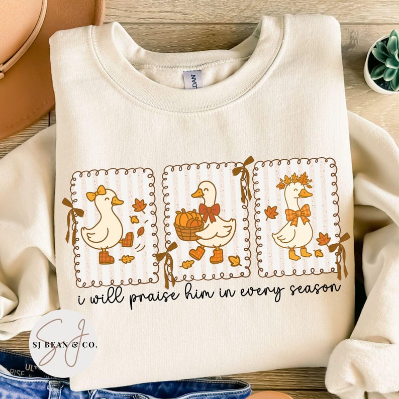 May include: Cream-colored sweatshirt featuring three cartoon ducks in decorative frames. Each duck is adorned with fall-themed accessories like bows, leaves, and pumpkins. The text "i will praise him in every season" is printed below the ducks.
