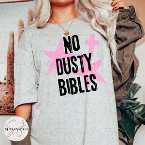 Peut inclure: Un t-shirt gris clair &agrave; manches courtes avec le texte noir NO DUSTY BIBLES imprim&eacute; dans une police us&eacute;e. Le motif comprend deux grandes &eacute;toiles roses et une petite croix rose positionn&eacute;es derri&egrave;re le lettrage noir en gras.