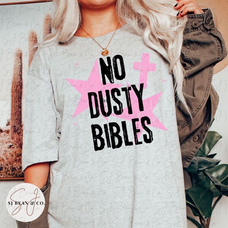 Peut inclure: Un t-shirt gris clair &agrave; manches courtes avec le texte noir NO DUSTY BIBLES imprim&eacute; dans une police us&eacute;e. Le motif comprend deux grandes &eacute;toiles roses et une petite croix rose positionn&eacute;es derri&egrave;re le lettrage noir en gras.