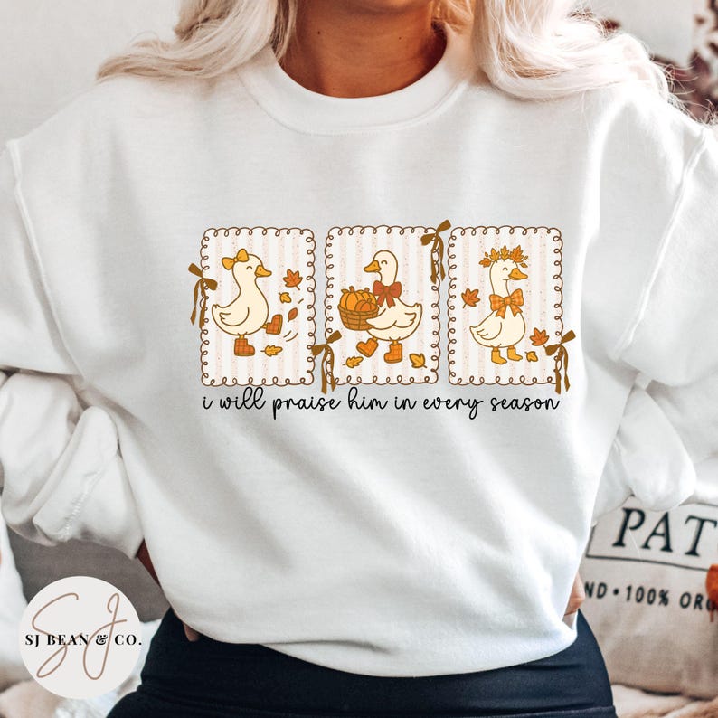 May include: White sweatshirt featuring a whimsical design of three ducks in seasonal settings. Each duck is within a framed design with fall leaves and bows. The text "i will praise him in every season" is printed below the ducks.