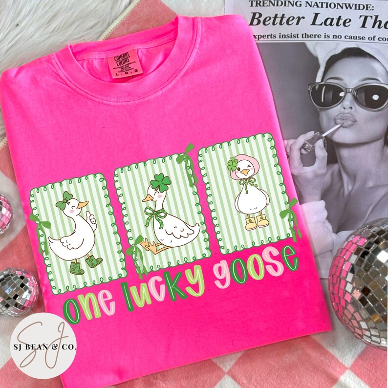 Puede incluir: Una camiseta rosa brillante con tres gansos de dibujos animados en marcos a rayas verdes y blancas, cada uno adornado con accesorios tem&aacute;ticos del D&iacute;a de San Patricio. El texto "one lucky goose" est&aacute; impreso en verde y rosa. La camiseta tiene una etiqueta Comfort Colors.