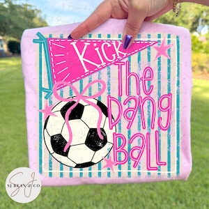 Bright pink text reading “Kick the dang ball” layered over a black and white soccer ball with a bow, set on a teal striped background with a pennant banner and star accents in a retro style.