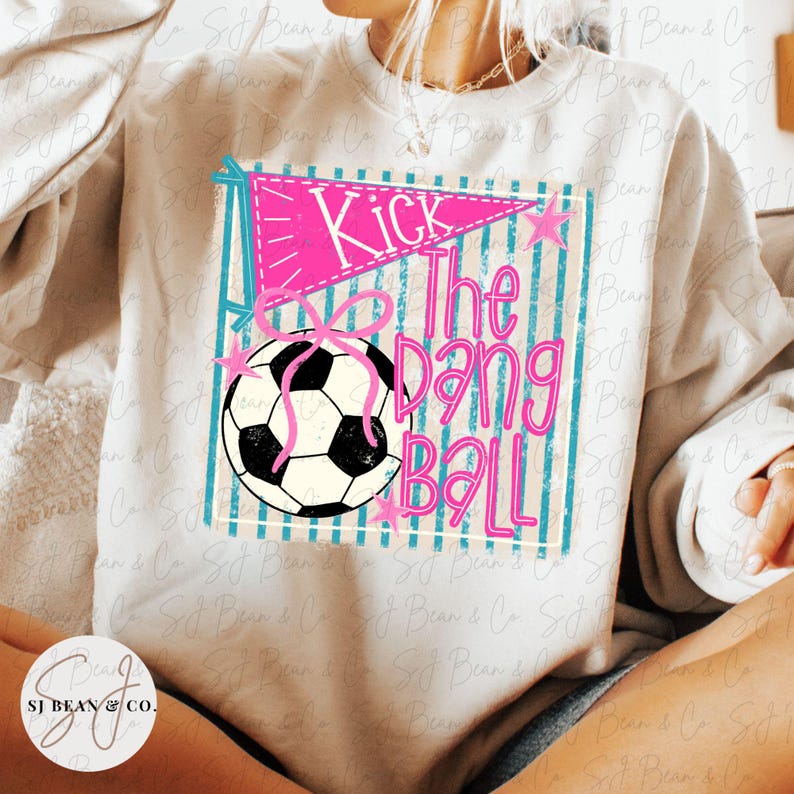 K&ouml;nnte beinhalten: Ein beiges Sweatshirt mit Rundhalsausschnitt und einem Grafikdruck eines schwarz-wei&szlig;en Fu&szlig;balls, eines rosa Wimpels und einer rosa Schleife. Der Text lautet Kick The Dang Ball in rosa Schreibschrift. Das Design enth&auml;lt t&uuml;rkise vertikale Streifen.