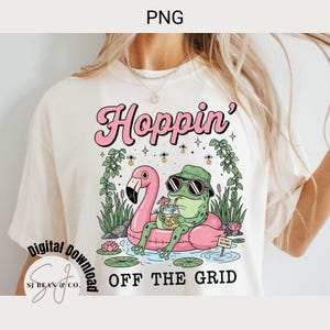May include: Cream-colored t-shirt with a graphic of a frog in sunglasses and a hat, relaxing on a pink flamingo float. The text "Hoppin' OFF THE GRID" is above and below the image. The design includes water lilies and foliage.