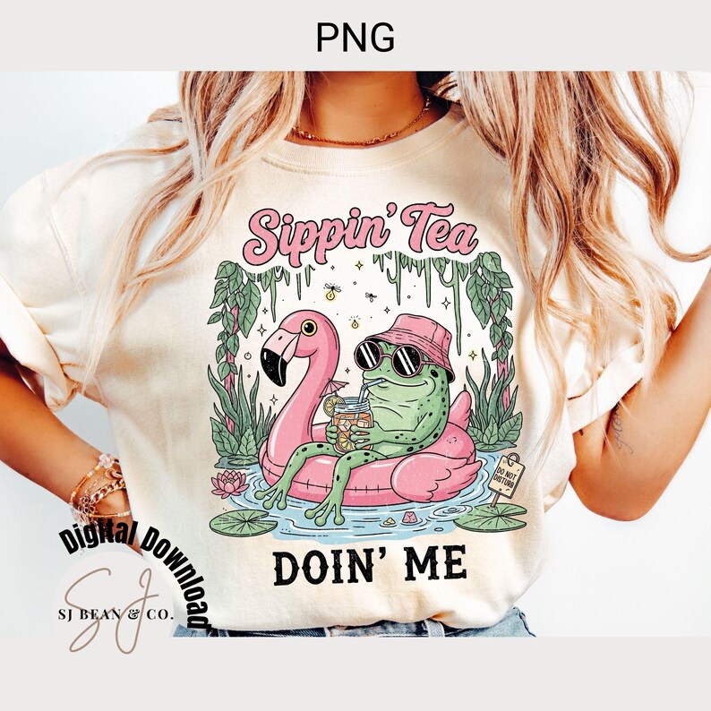 以下が含まれることがあります： クリーム色のTシャツ。サングラスをかけたカエルがピンクのフラミンゴの浮き輪に乗っているグラフィック。カエルは瓶から飲んでいる。テキストは「Sippin' Tea」と「Doin' Me」。デザインは沼地のシーンに囲まれています。
