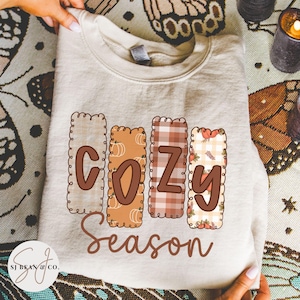 May include: A beige sweatshirt with the words "COZY Season" in a fall-themed design. The letters are outlined with a decorative border and filled with patterns including pumpkins and plaid. The sweatshirt is held by hands.
