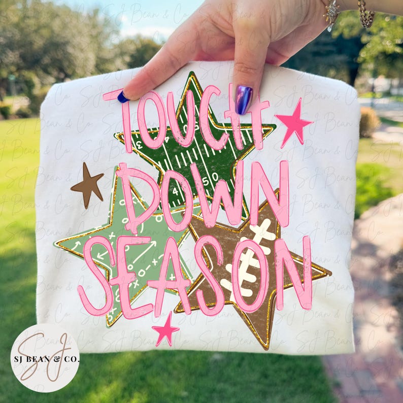 May include: A white t-shirt featuring a graphic of three overlapping stars in dark green, light green, and brown. The stars contain football field markings and play diagrams. The text "TOUCH DOWN SEASON" is printed in large, pink, cursive letters.
