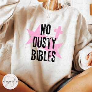 Peut inclure: Un sweat-shirt &agrave; col rond de couleur cr&egrave;me arborant un motif graphique avec le texte noir NO DUSTY BIBLES. Le design comprend une &eacute;toile rose et un symbole de genre f&eacute;minin rose, avec de petites croix roses entourant le lettrage central.