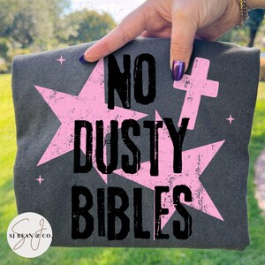 Peut inclure: Un t-shirt gris anthracite avec une impression graphique vieillie portant le texte noir NO DUSTY BIBLES. Le motif comprend de grandes &eacute;toiles rose clair et une petite croix rose, agr&eacute;ment&eacute;es de minuscules &eacute;toiles &agrave; quatre branches.