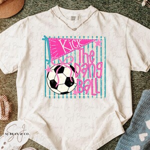 K&ouml;nnte beinhalten: Wei&szlig;es T-Shirt mit einem Aufdruck &bdquo;Kick The Dang Ball&ldquo;. Die Grafik enth&auml;lt einen rosa Wimpel, einen Fu&szlig;ball und t&uuml;rkisfarbene Streifen. Das Shirt ist von der Marke Comfort Colors.