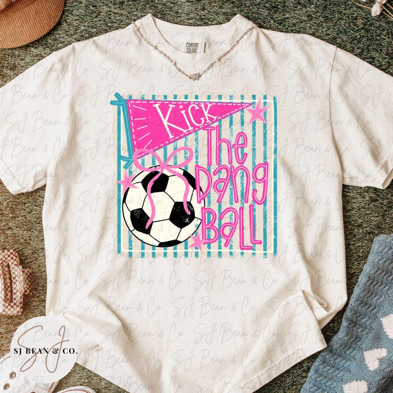 K&ouml;nnte beinhalten: Wei&szlig;es T-Shirt mit einem Aufdruck &bdquo;Kick The Dang Ball&ldquo;. Die Grafik enth&auml;lt einen rosa Wimpel, einen Fu&szlig;ball und t&uuml;rkisfarbene Streifen. Das Shirt ist von der Marke Comfort Colors.