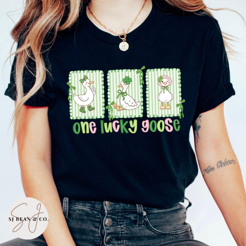 Puede incluir: Camiseta negra con un gr&aacute;fico de tres gansos blancos, cada uno en un marco a rayas verdes, con detalles verdes y rosas. El texto "one lucky goose" est&aacute; debajo de los gansos en verde y rosa. Una prenda casual.
