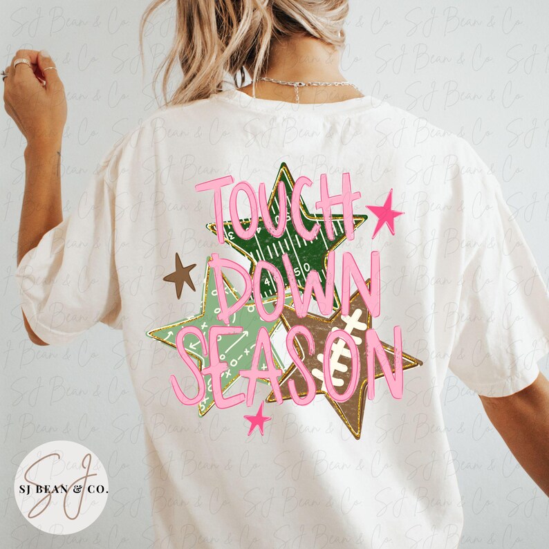May include: An off-white t-shirt featuring a back graphic of three stars in shades of green and brown, decorated with football field patterns and the pink text "TOUCH DOWN SEASON" layered over the stars, accompanied by small pink and brown stars.