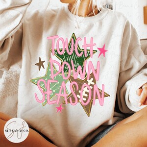 May include: A cream-colored crewneck sweatshirt featuring a graphic of three overlapping stars in green and brown, decorated with football field patterns and a football lace design. The text TOUCH DOWN SEASON is printed in large, bright pink letters.