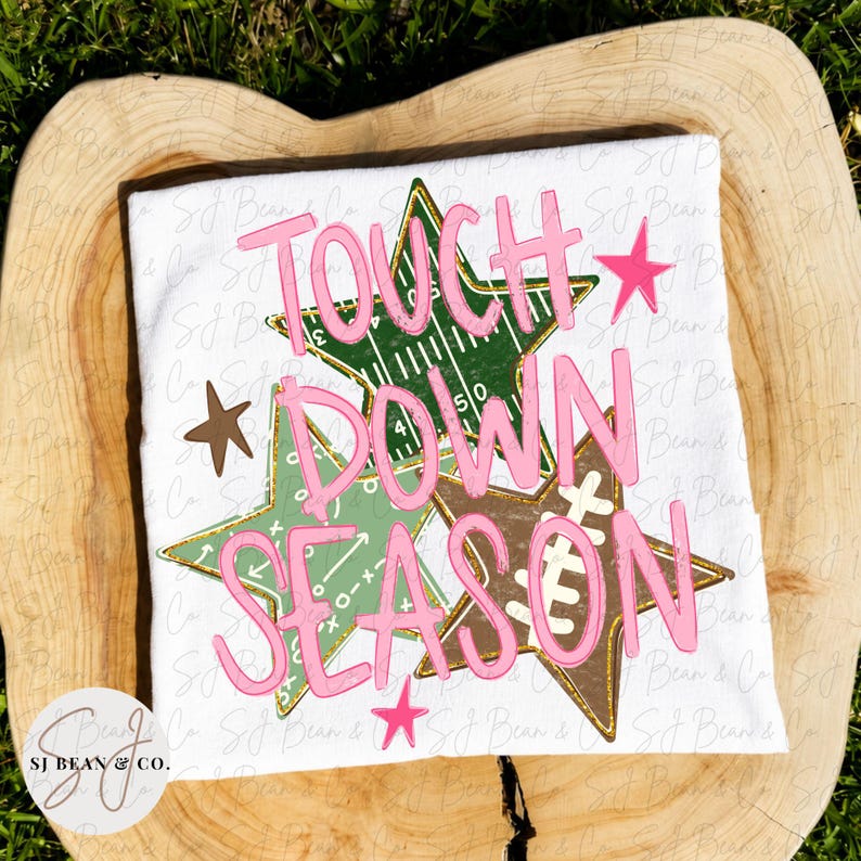 May include: A white t-shirt featuring the pink text TOUCH DOWN SEASON layered over three stars. One star has a green football field pattern, another has white play diagrams, and the third has a football lace design. Small pink and brown stars surround.