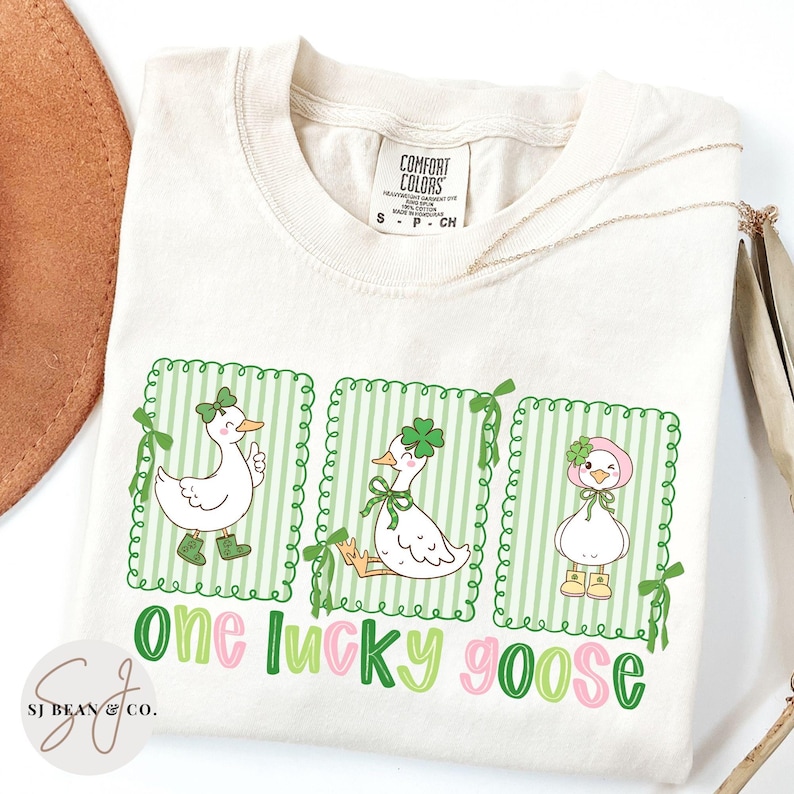 Puede incluir: Camiseta color crema con tres gansos de dibujos animados en marcos a rayas verdes, cada uno adornado con accesorios del D&iacute;a de San Patricio. El texto "one lucky goose" est&aacute; escrito en verde y rosa. La camiseta tiene una etiqueta Comfort Colors.
