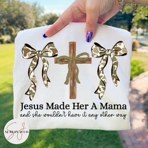 May include: White t-shirt featuring a cross and two bows with a camouflage pattern. The text "Jesus Made Her A Mama and she wouldn't have it any other way" is printed below the design. The brand logo is in the bottom left corner.