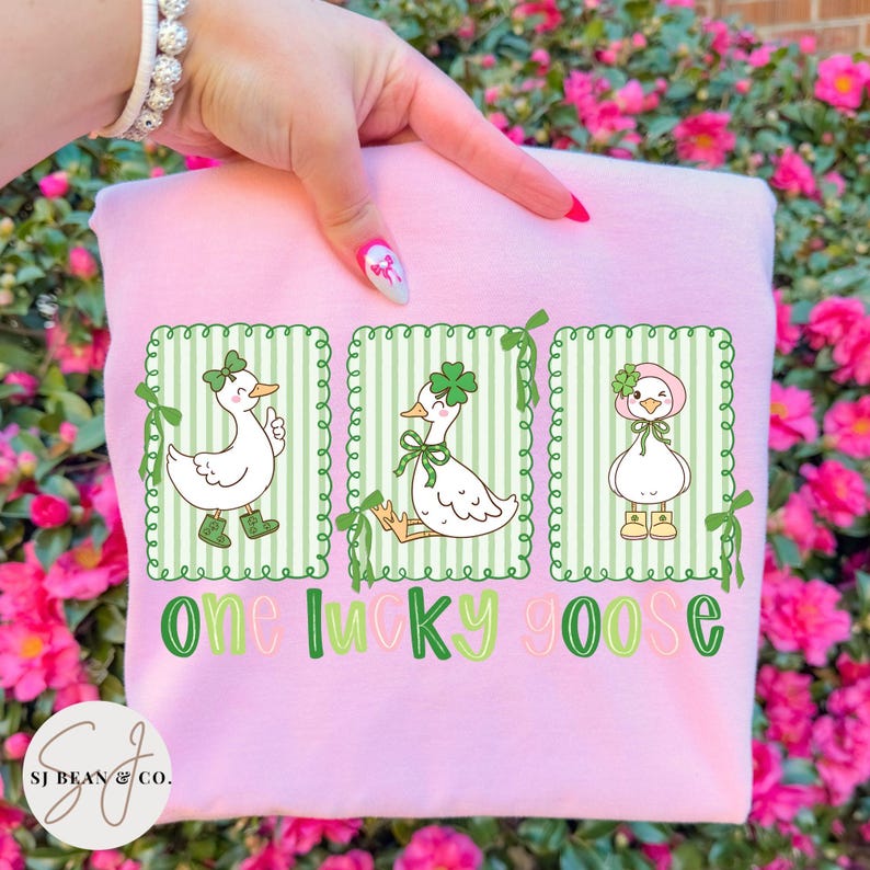 Puede incluir: Sudadera rosa claro con un dise&ntilde;o del D&iacute;a de San Patricio. Presenta tres gansos de dibujos animados en rect&aacute;ngulos con marco verde, cada uno con tr&eacute;boles y detalles verdes. El texto "One Lucky Goose" est&aacute; impreso en verde debajo.