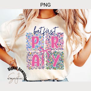 Könnte beinhalten: Ein weißes T-Shirt mit dem Text "but first pray" in rosa Buchstaben, umgeben von einem Blumenmuster. Die Buchstaben befinden sich in einer Kastenform mit einer rosa Schleife in der Mitte.