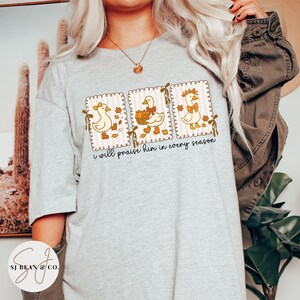 May include: A light gray t-shirt featuring three framed illustrations of cartoon ducks. Each duck is decorated with fall-themed elements. The text "I will praise him in every season" is printed below the illustrations. The shirt has a crew neck.