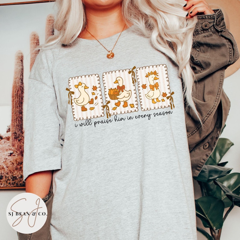 May include: A light gray t-shirt featuring three framed illustrations of cartoon ducks. Each duck is decorated with fall-themed elements. The text "I will praise him in every season" is printed below the illustrations. The shirt has a crew neck.