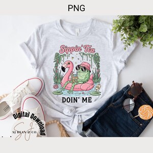 以下が含まれることがあります： 「Sippin' Tea Doin' Me」の文字と、サングラスをかけたカエルのイラストが描かれたライトグレーのTシャツ。カエルはピンクのフラミンゴの浮き輪でくつろいでいます。カエルは飲み物を持っています。白いスニーカーとブルージーンズも写っています。