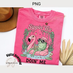 以下が含まれることがあります： 「Sippin' Tea Doin' Me」の文字と、サングラスと帽子をかけたカエルがフラミンゴの浮き輪でくつろいでいるグラフィックが描かれたピンク色のTシャツ。カエルは飲み物を持っています。シャツには「Comfort Colors」の文字がタグにあります。
