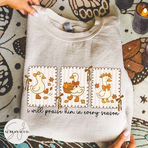 May include: Beige sweatshirt featuring three cartoon ducks in framed designs. Each duck has a bow and is surrounded by fall leaves. The text "i will praise him in every season" is printed below the ducks. The sweatshirt is held by a person.