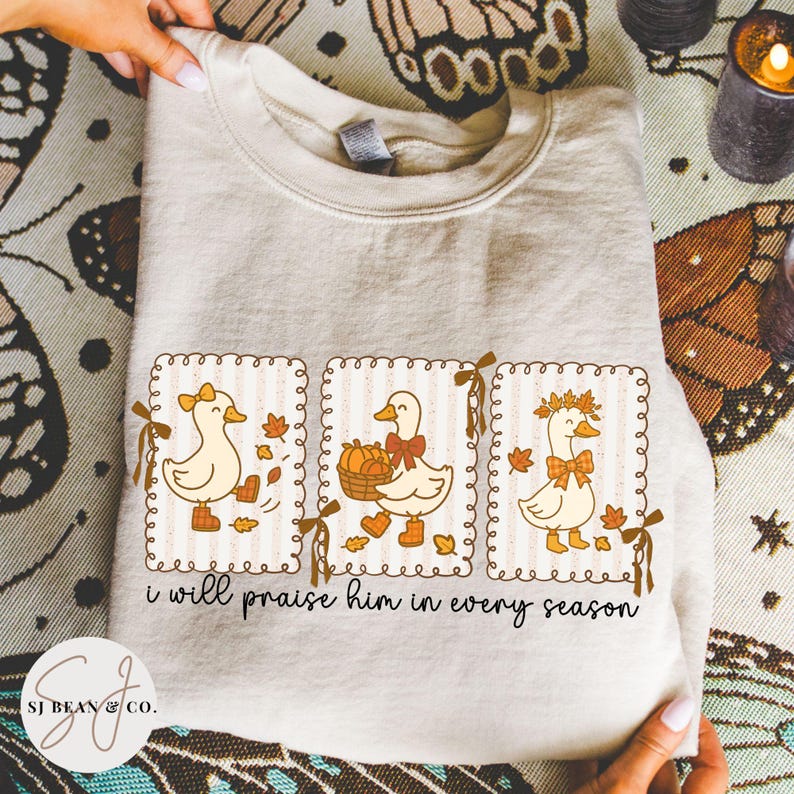 May include: Beige sweatshirt featuring three cartoon ducks in framed designs. Each duck has a bow and is surrounded by fall leaves. The text "i will praise him in every season" is printed below the ducks. The sweatshirt is held by a person.