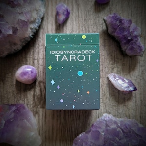 May include: A tarot card deck box with the text "IDIOSYNCRADECK TAROT" on a dark teal background with colourful star and dot graphics. The box is surrounded by amethyst crystals and stones on a wooden surface.