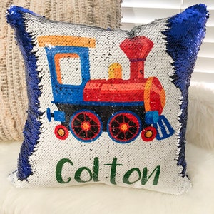 May include: A white and blue reversible sequin pillow with a colorful train and the name "Colton" printed on it. The pillow is decorated with blue sequins on one side and white sequins on the other side.