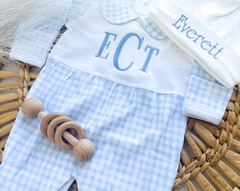 May include: A light blue and white gingham footed sleeper with a white collar and chest panel embroidered with the letters ECT in blue thread. A matching white beanie with the name Everett embroidered in blue and a wooden ring rattle are included.