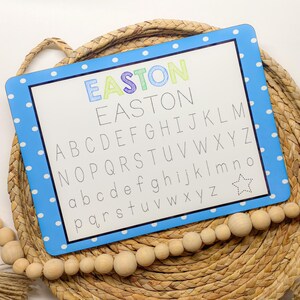 May include: A blue and white dry erase board with a polka dot border. The board has the name Easton written in rainbow colors at the top and the alphabet written in black below. The board is sitting on a woven basket.