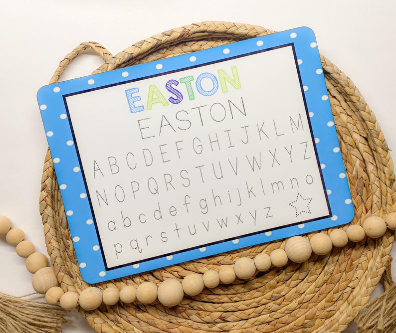May include: A blue and white dry erase board with a polka dot border. The board has the name Easton written in rainbow colors at the top and the alphabet written in black below. The board is sitting on a woven basket.
