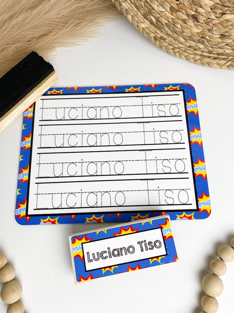 May include: A blue and yellow dry-erase board with a superhero theme. The board has four lines for writing practice. The name "Luciano Tiso" is written on the board. A separate wooden block with the same superhero theme and the name "Luciano Tiso" is also shown.