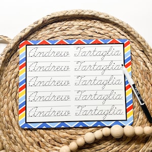 May include: A white dry-erase board with blue, red, and yellow stripes on the edges. The board has five lines of cursive handwriting that say "Andrew Fartaglia" in black ink.