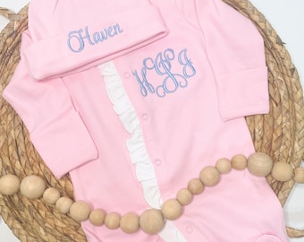 May include: Pink baby footed sleeper and matching hat set. The sleeper features a white ruffle down the front and light blue embroidered initials. The hat is embroidered with the name "Haven" in light blue. The set is displayed on a woven basket.